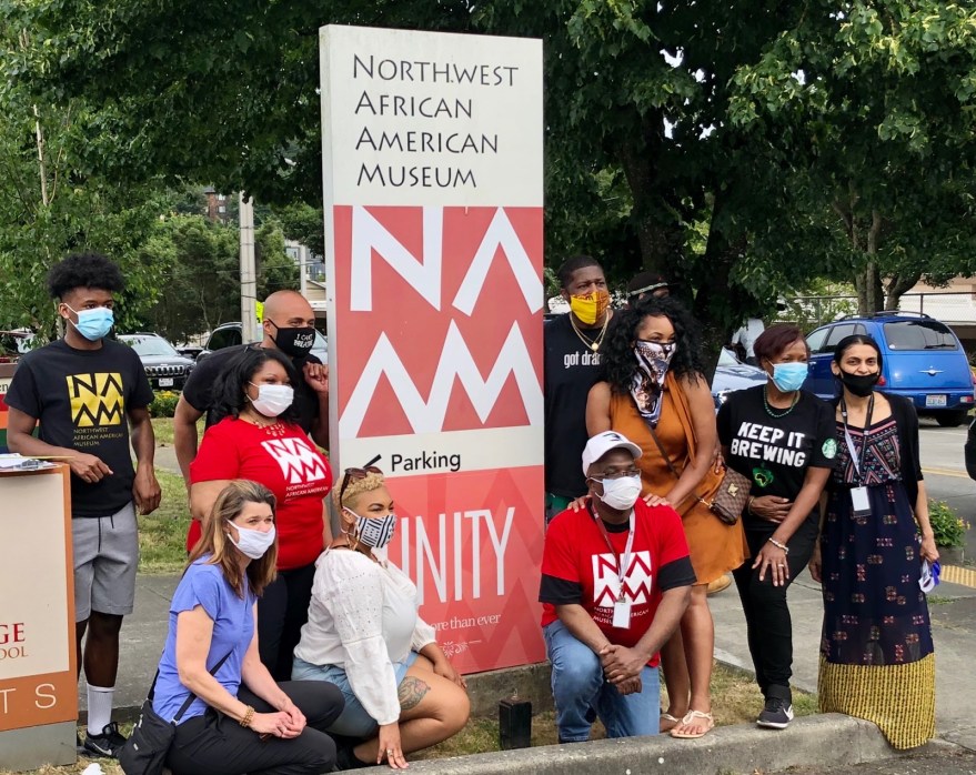 A group of people wearing masks and t-shirts with the museum's logo in front of a sign for the museum in a parking lot.