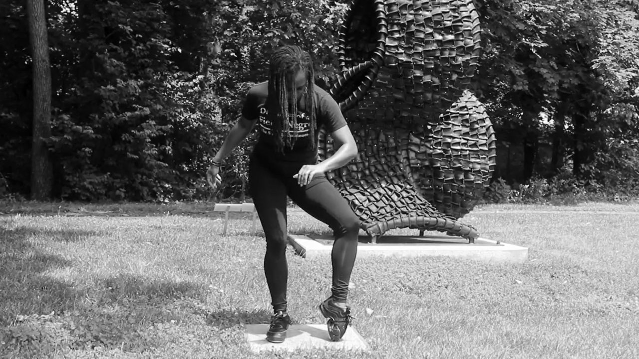 A person bent over in a dance move in front of a black undulating sculpture in an outdoor setting