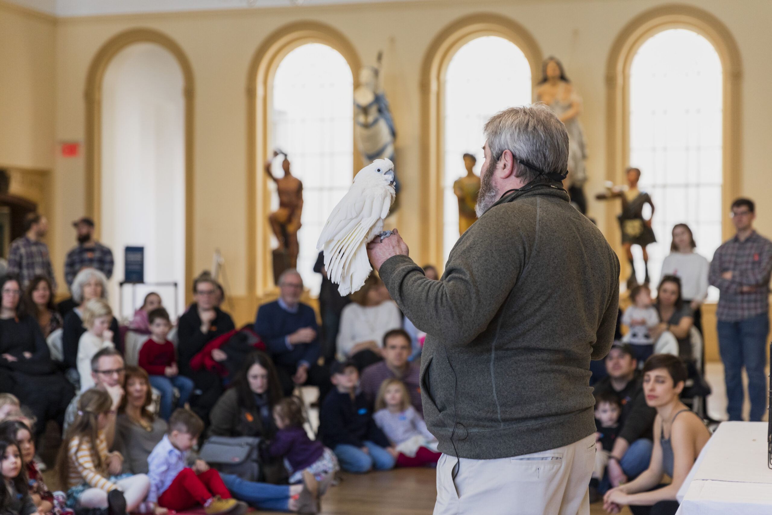 A speaker addressing an audience inside a museum with a bird perched on his hand