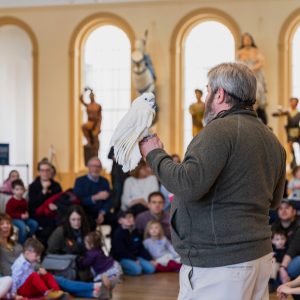 A speaker addressing an audience inside a museum with a bird perched on his hand