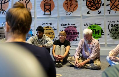 A group of people seated in a circle on the floor in front of drawings hanging on a gallery wall