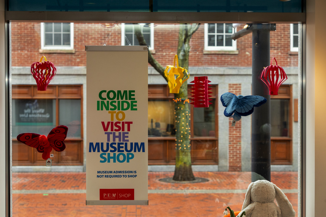 A sign on a window reading "Come inside to visit the museum shop" in rainbow-colored font, with colorful paper lanterns and stuffed butterflies hanging around it