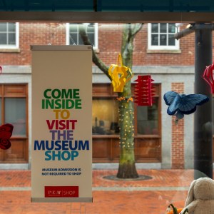 A sign on a window reading "Come inside to visit the museum shop" in rainbow-colored font, with colorful paper lanterns and stuffed butterflies hanging around it