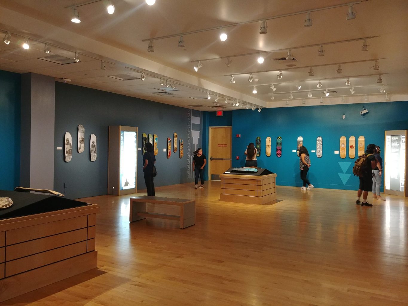 Students wearing masks inside of a museum gallery looking at skateboard decks mounted on the wall