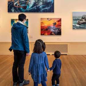 One adult and two children visitors looking at a gallery wall of paintings depicting fantastical shipwrecks