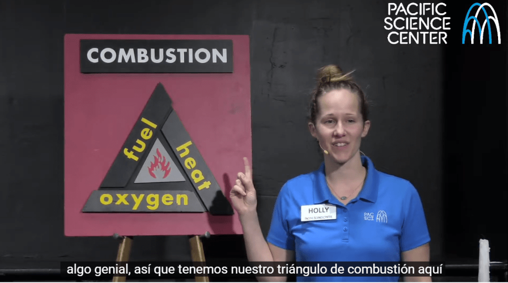 An educator leading a program in front of a poster reading "Combustion: fuel, heat, oxygen" with Spanish subtitles on the screen below