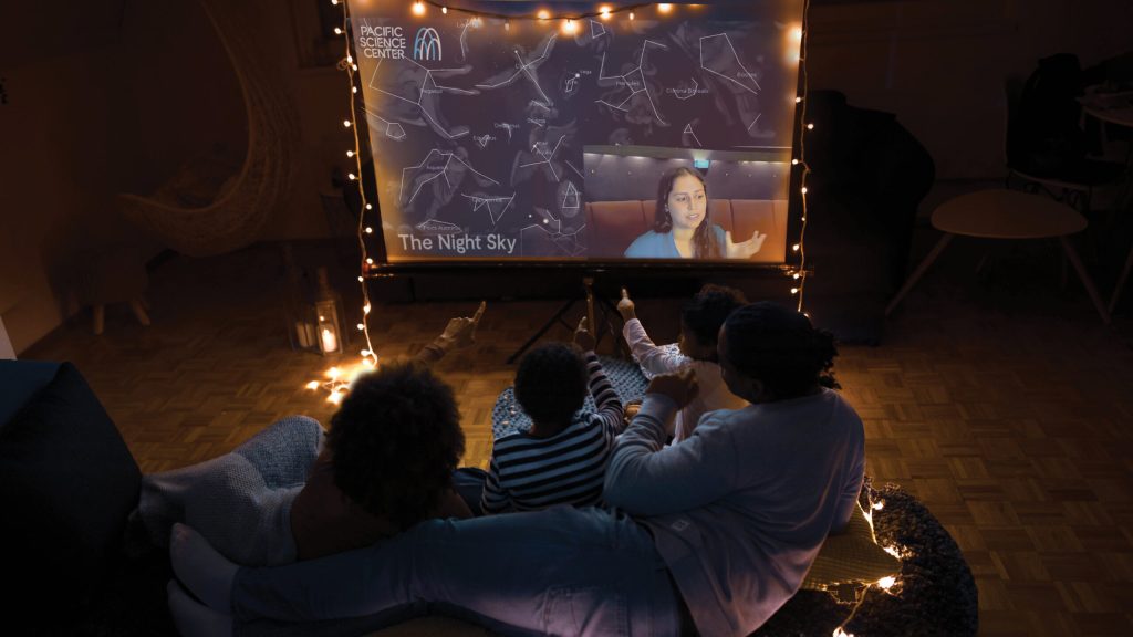A group of small children and adults watching a Pacific Science Center virtual program on a projector in a darkened room with fairy lights.