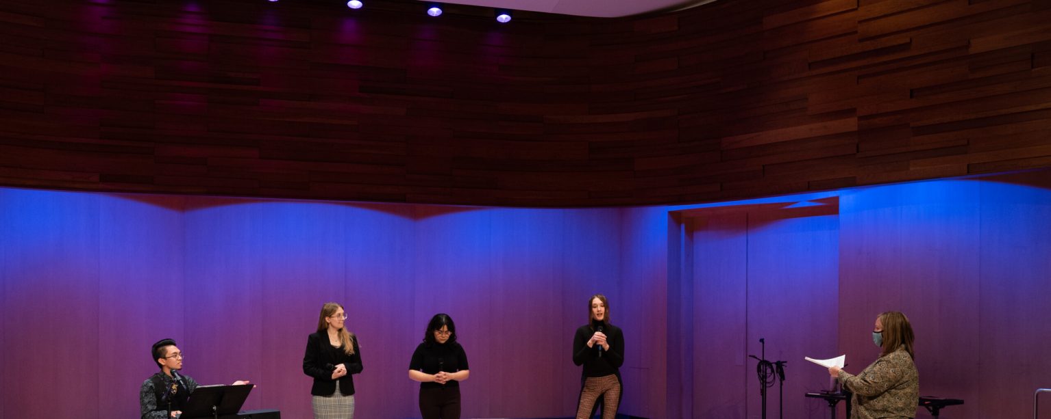 A group of performers standing on a stage with a director reading from notes to them