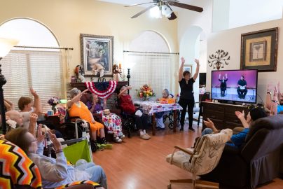 A group of seniors and senior care staff seated around a television showing a video of a drummer and instructor