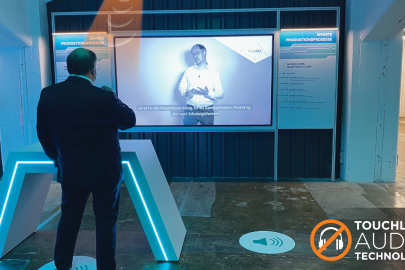 A gallery visitor standing on a floor decal with a volume icon, with a graphic insert of a beam extending down to their head, and a video playing on the screen they face.