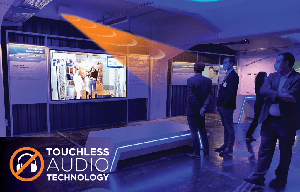 A group of gallery visitors watching a video on a screen with a graphic of a light beam extending onto them and the text "Touchless Audio Technology"