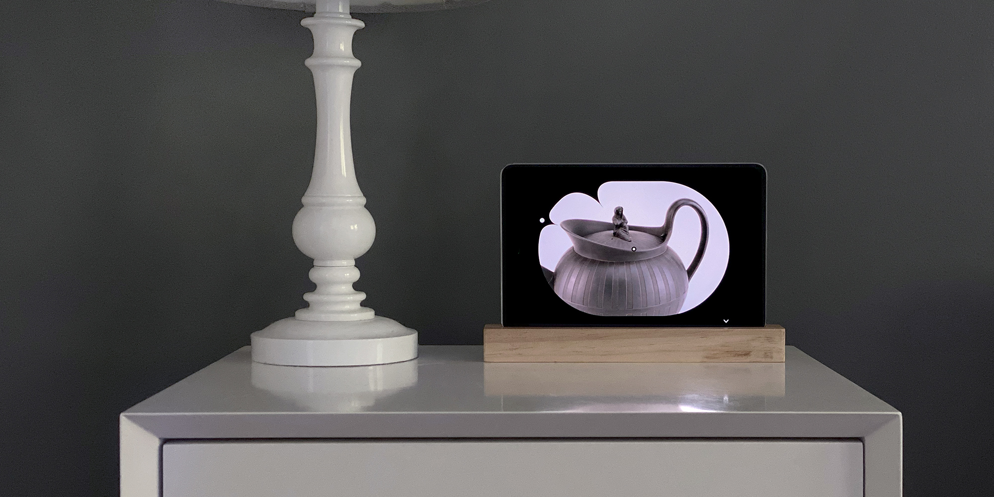 A rendering of a bedside table with a digital screen on a pedestal showing a picture of a teapot with a minimalist clock shape aligned with the teapot's spot