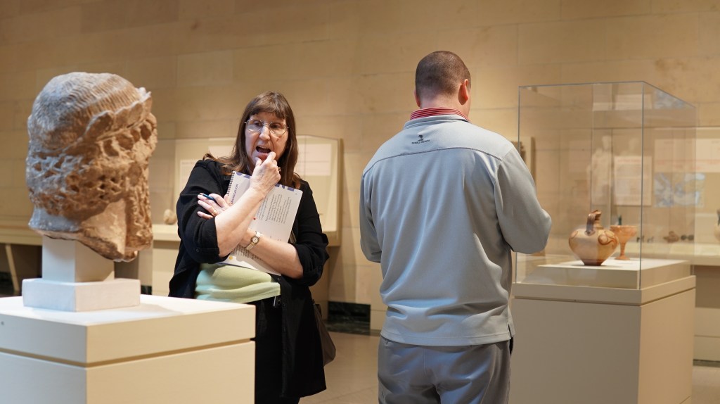 Two workshop participants study sculpture and pottery in a museum gallery while holding activity books