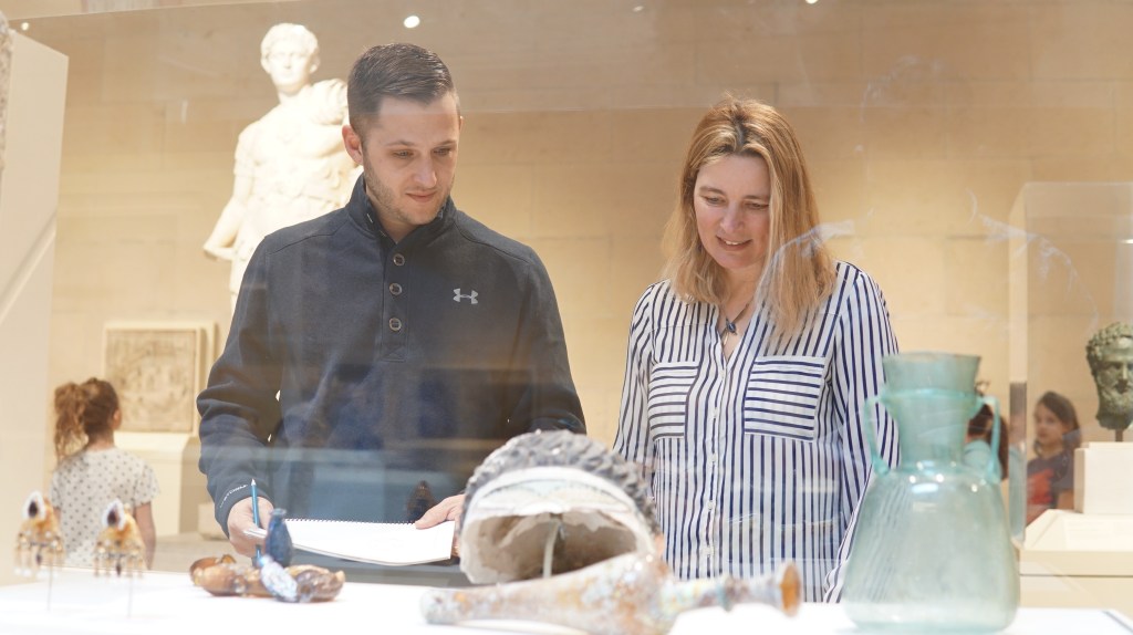 Two workshop participants look at works of sculpture and pottery inside a vitrine while holding activity books