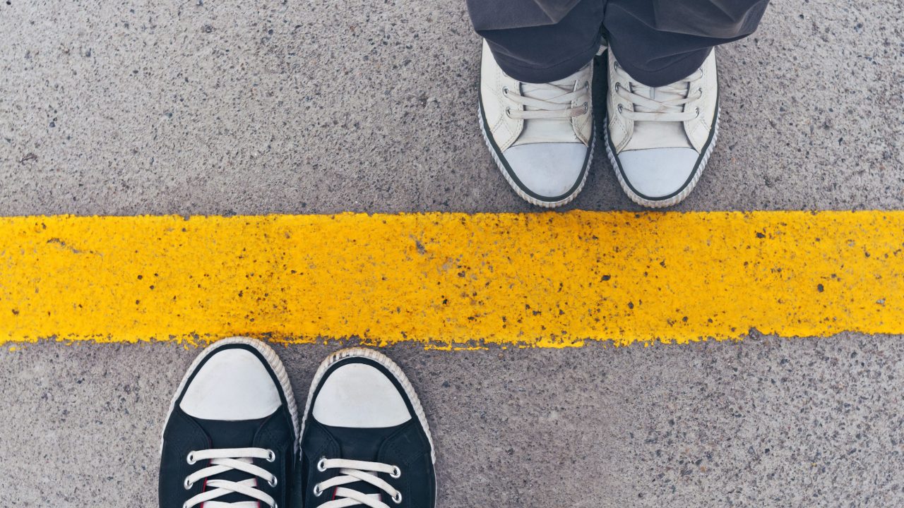 Two people's shoes seen from above standing on the opposite sides of a yellow line