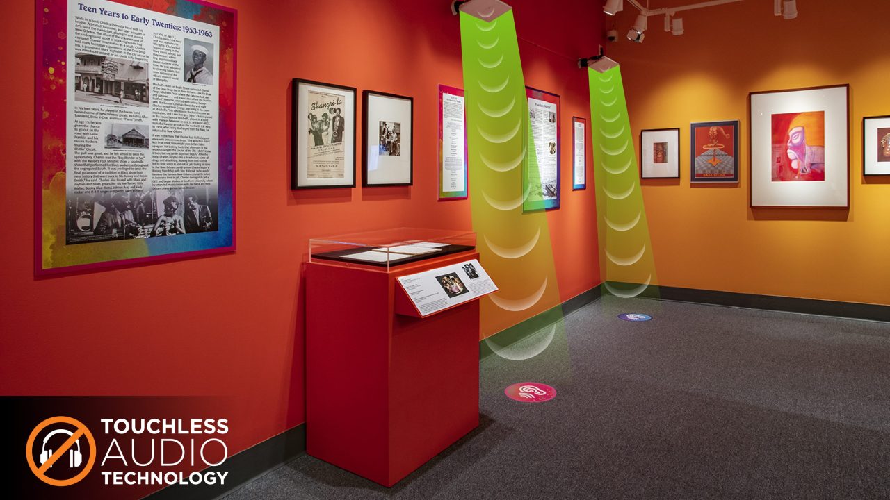 A museum gallery displaying music ephemera with two speakers hanging over floor decals where visitors can stand to trigger audio