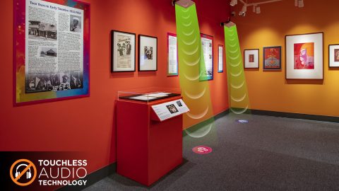 A museum gallery displaying music ephemera with two speakers hanging over floor decals where visitors can stand to trigger audio