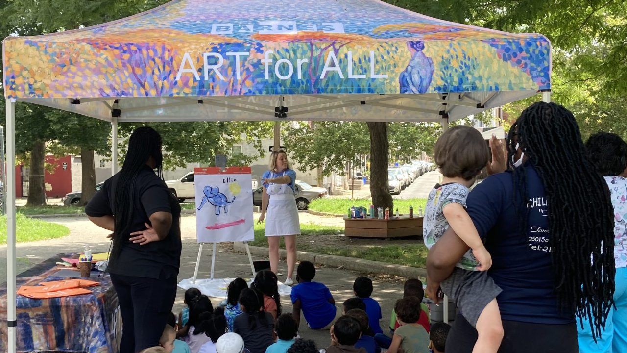 A group of children and caregivers watching a storytime taking place under a tent labeled "Barnes / Art for All"
