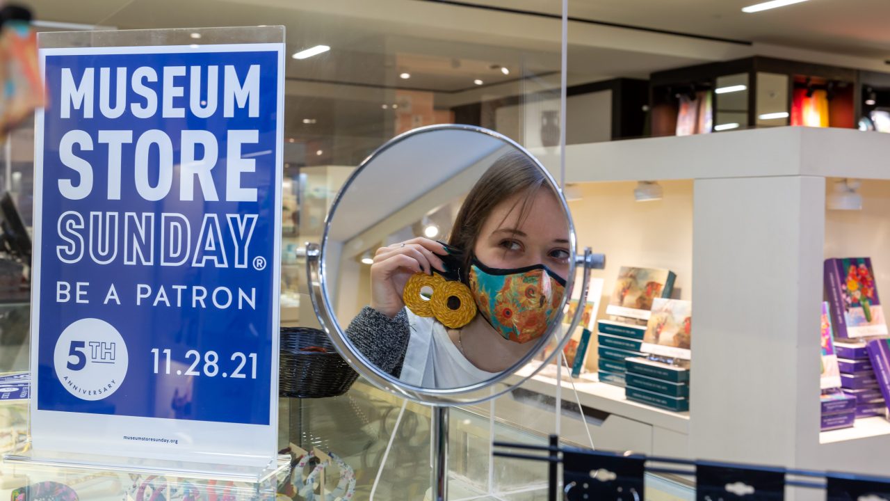 A shopper looking at sunflower-shaped earrings in a mirror in a museum shop next to a sign for Museum Store Sunday