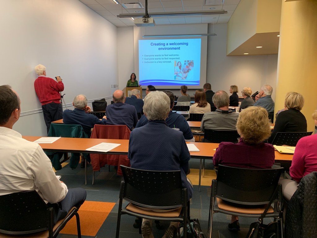 A group of people watching a training, where the presenter is showing a presentation slide labeled "Creating a welcoming environment."