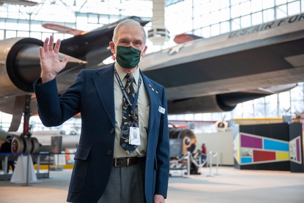 A volunteer standing in a museum space wearing a mask and waving