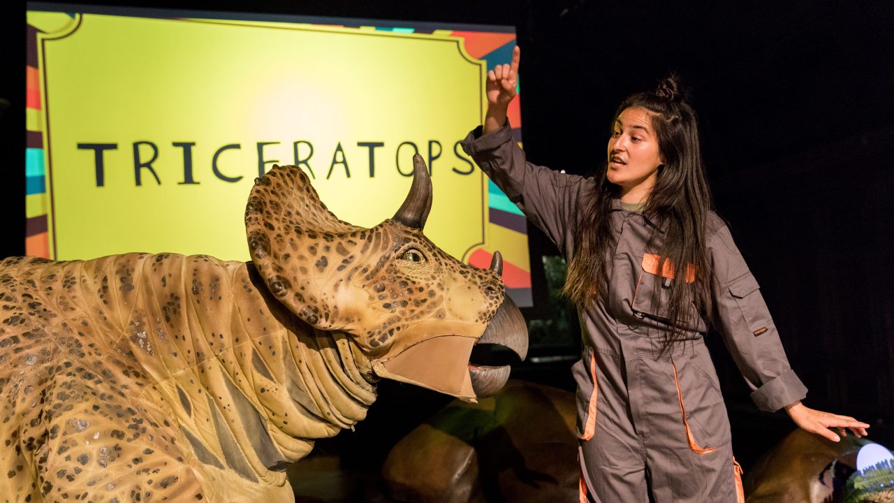A performer wearing a jumpsuit gestures in front of a performer inside of a realistic dinosaur costume
