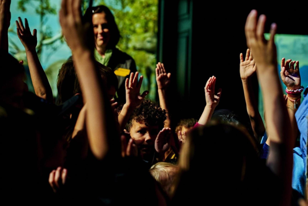 A performer looks out into an audience of children raising their hands