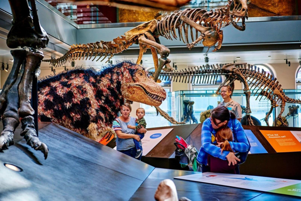 Visitors standing in a display of dinosaur skeletons and looking at a lifelike dinosaur puppet