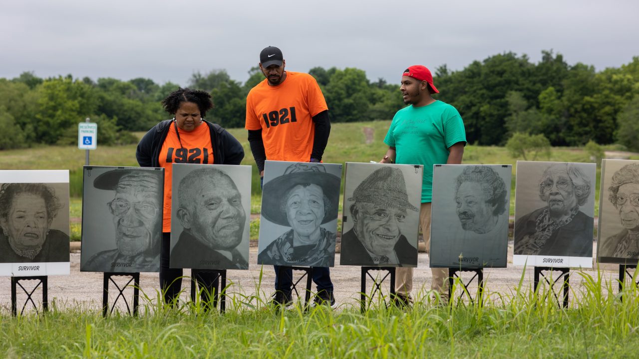 A group of three people looking at black-and-white portraits of people labeled "survivor" in an outdoor setting