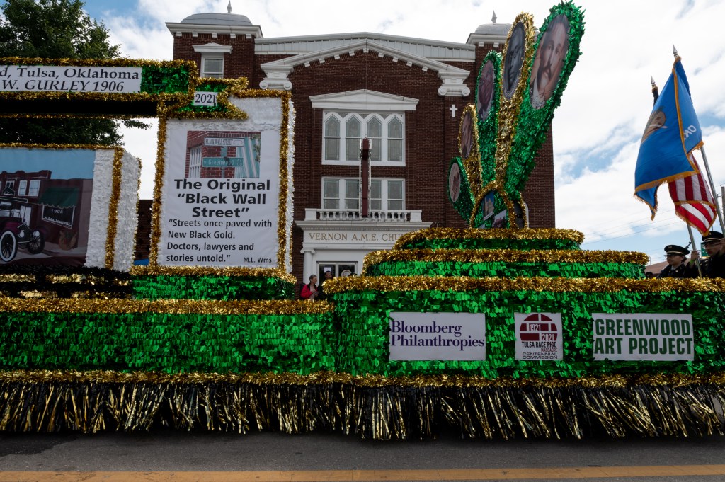 A parade float with the logos of Bloomberg Philanthopies and the Greenwood Art Project and the text "The Original 'Black Wall Street' 'Streets once paved with New Black Gold. Doctors, lawyers, and stories untold."