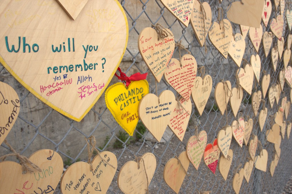 A big plywood heart on a chainlink fence reading "Who will you remember?" surrounded by smaller plywood hearts with messages like "Philando Castile."