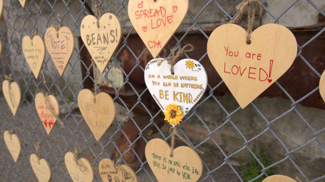 Plywood hearts attached to a chainlink fence and decorated with messages like "You are LOVE" and "In a world where you can be anything, be kind."
