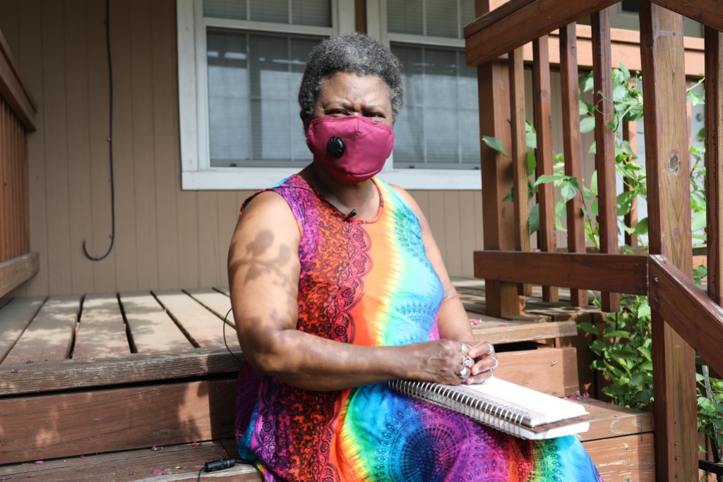 A person wearing a rainbow-colored dress and mask and holding an open notebook on a porch