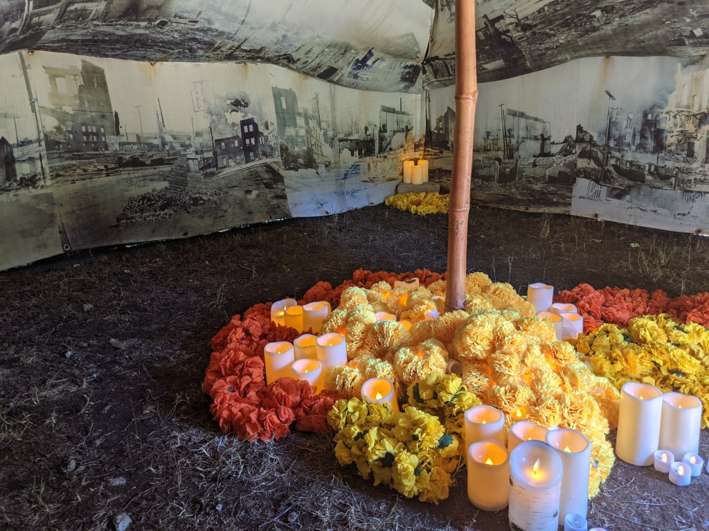 The inside of a tent screenprinted with images of burned buildings, with pillar candles and cut flowers organized into bundles on the ground