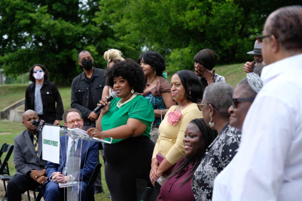 A speaker surrounded by a group of people at a podium with the logo for the Greenwood Art Project