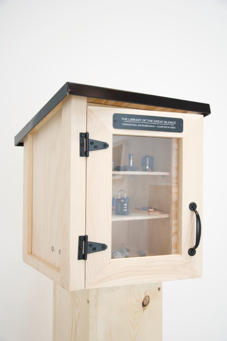 A wooden cabinet on a pedestal modeled after a "Little Free Library" with a glass pane on the front to see into the shelves from the outside. A plaque on the front reads "The Library of the Great Silence / A universal repository for interstellar research on planetary futures / Terrestrial microbranch charter no. 0001"