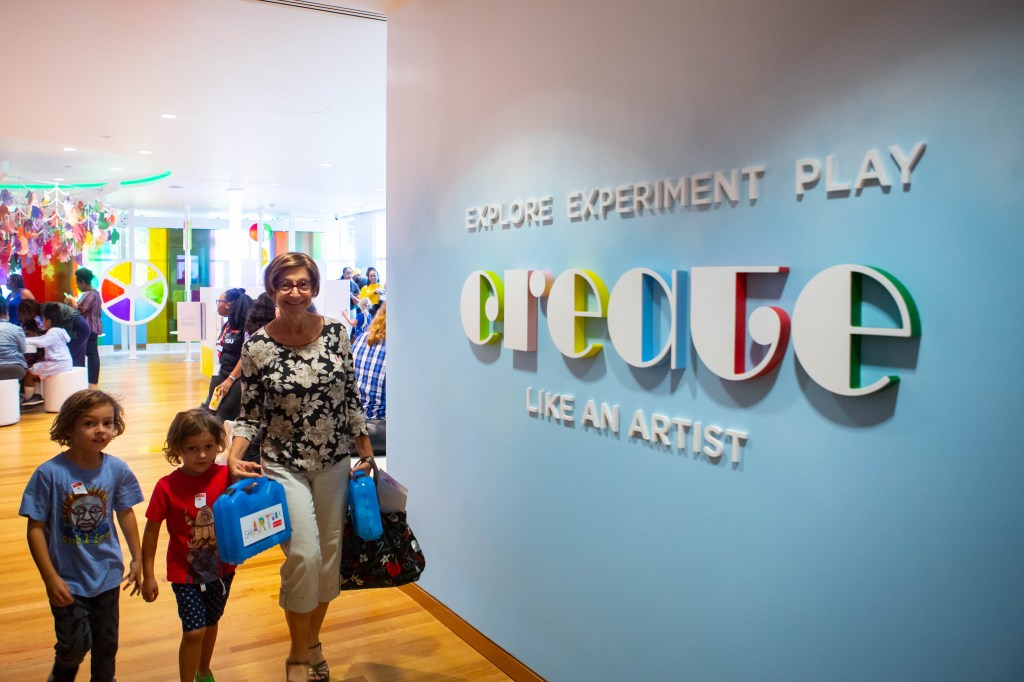 An adult guardian leading two children into a space with a sign reading "Explore, Experiment, Play: Create Like An Artist"
