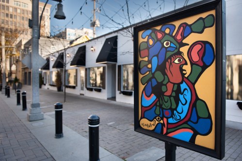 A reproduction of a stylized, brightly colored painting of a shaman on a pedestal bolted into the sidewalk on a commercial urban street