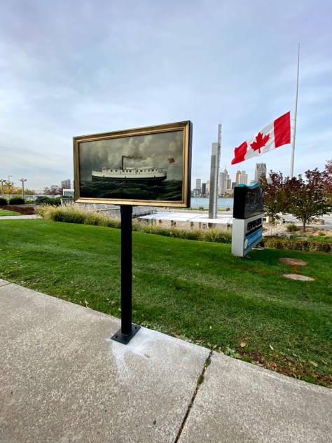 A reproduction of an oil painting of a ship on a pedestal bolted in the sidewalk with a view of an urban downtown in the background