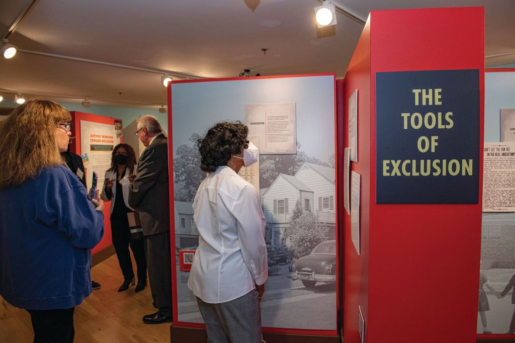 A number of people mill around an exhibition and one person stands in front of a particular group of artifacts on a wall. 