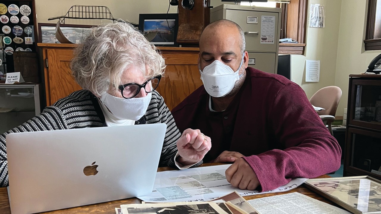 Two people sit with a sheet of paper and a laptop computer on a table in front of them.