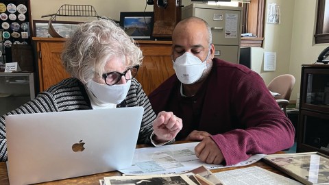 Two people sit with a sheet of paper and a laptop computer on a table in front of them.