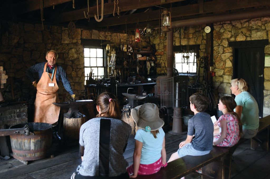 A person in a blacksmith apron stands in front of a family group explaining the blacksmith process.
