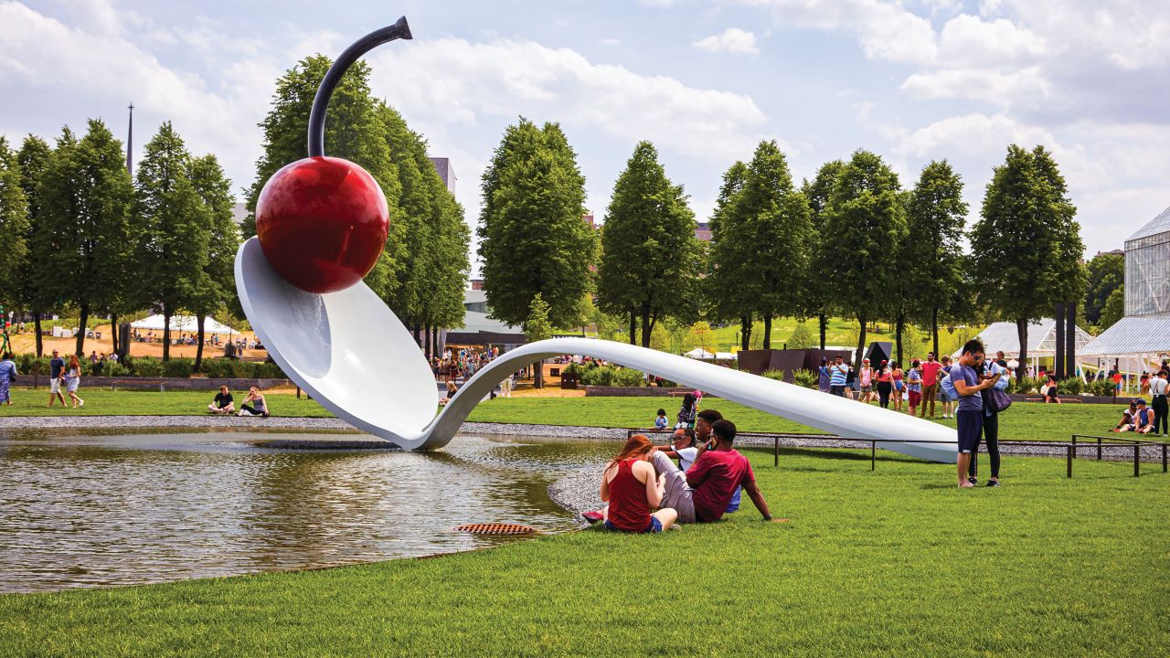 A sculpture of a giant spoon with a cherry sitting in the bowl lays over a small lake with people milling about on a lawn of green grass.