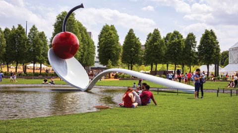 A sculpture of a giant spoon with a cherry sitting in the bowl lays over a small lake with people milling about on a lawn of green grass.