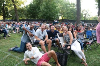 A group of people smile at the camera in a larger crowd of people viewing a concert on a lawn. 
