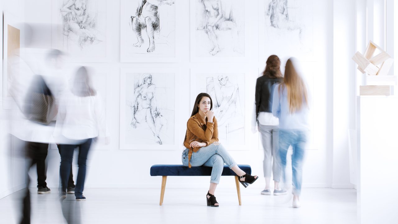 A person sitting on a bench in a museum gallery with people blurred out standing around them.