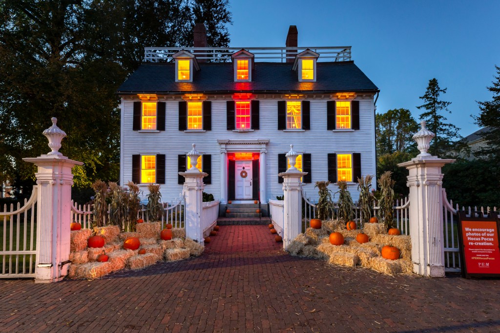 A historic house at sunset with pumpkins arranged outside the fence and lights on in the windows