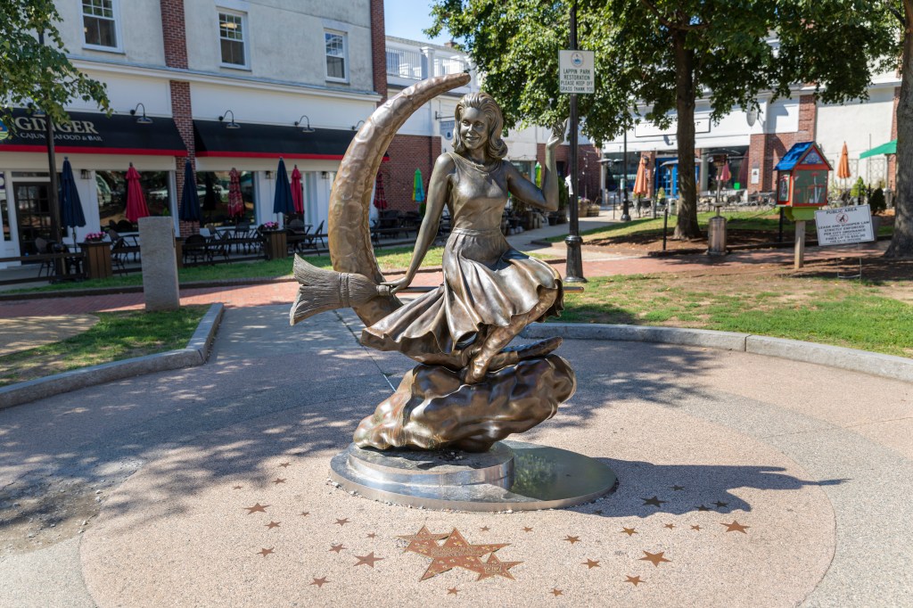 A bronze outdoor statue of a witch riding a broom on top of a crescent mood and cloud