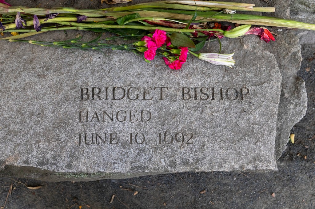 A gravestone reading "Bridget Bishop / Hanged / June 10 1692" with flowers on top of it
